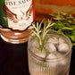 A glass of gin cocktail with ice and a rosemary sprig in the foreground, with a bottle of Five Saints Gin in the background on a dark surface.