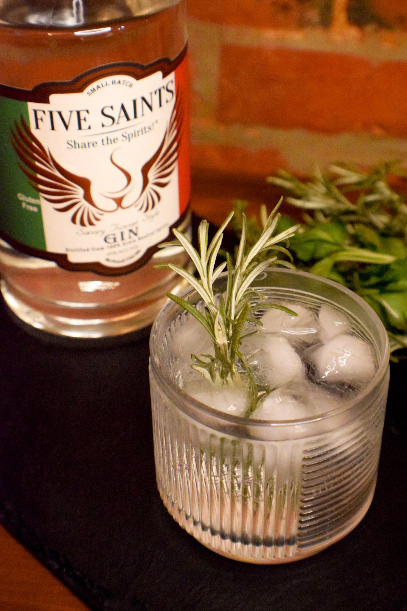 A glass of gin cocktail with ice and a rosemary sprig in the foreground, with a bottle of Five Saints Gin in the background on a dark surface.