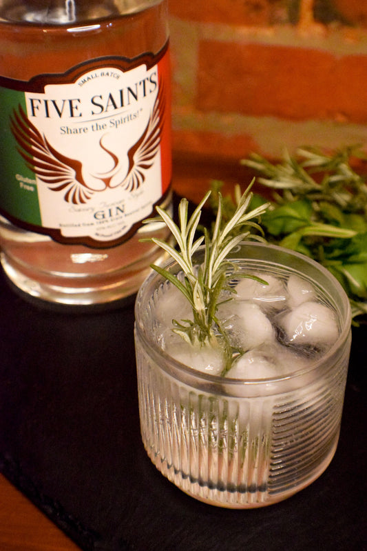 A glass of gin cocktail with ice and a rosemary sprig in the foreground, with a bottle of Five Saints Gin in the background on a dark surface.