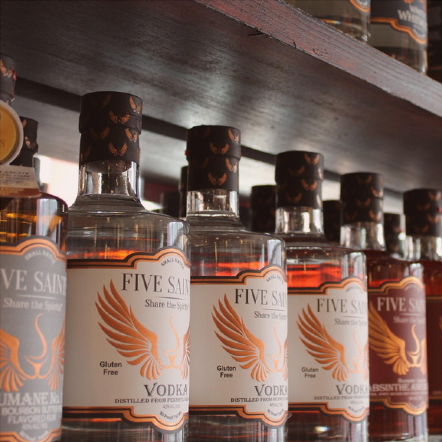 Multiple Five Saints Distilling spirit bottles lined up on a wooden shelf, including Triple Slow Distilled Vodka and other small batch spirits