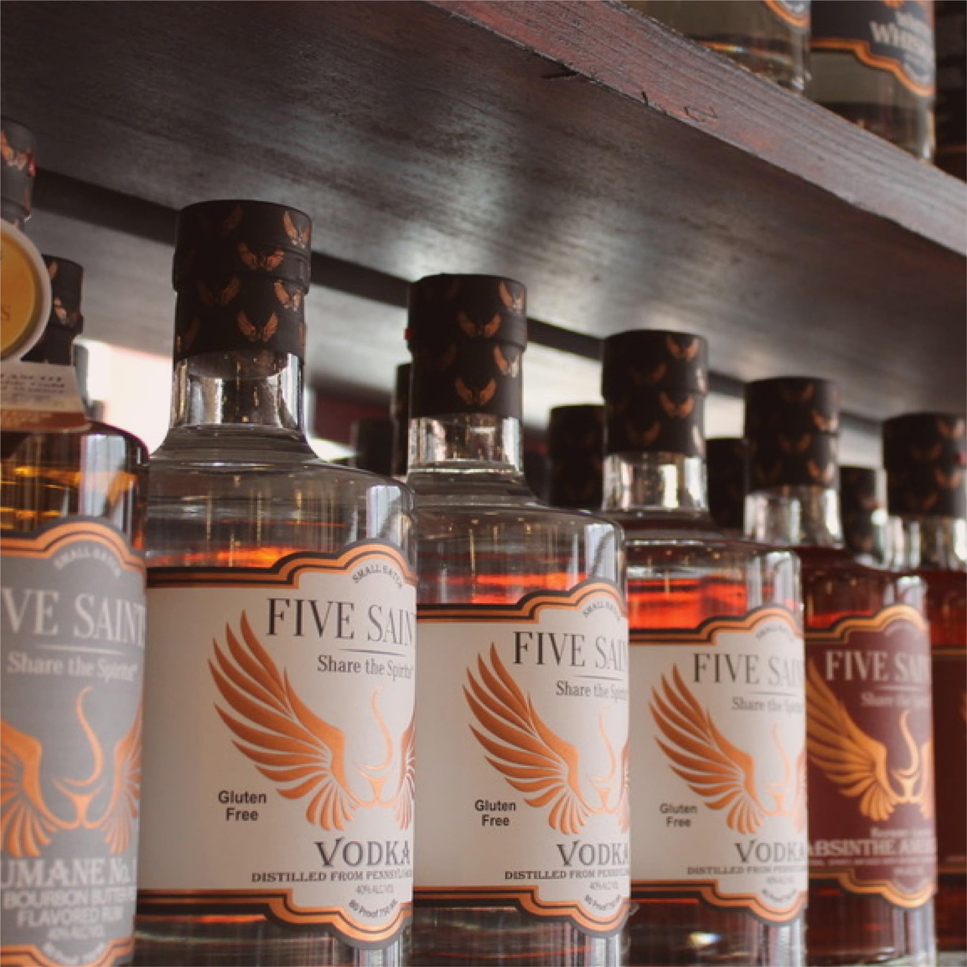 Multiple Five Saints Distilling spirit bottles lined up on a wooden shelf, including Triple Slow Distilled Vodka and other small batch spirits