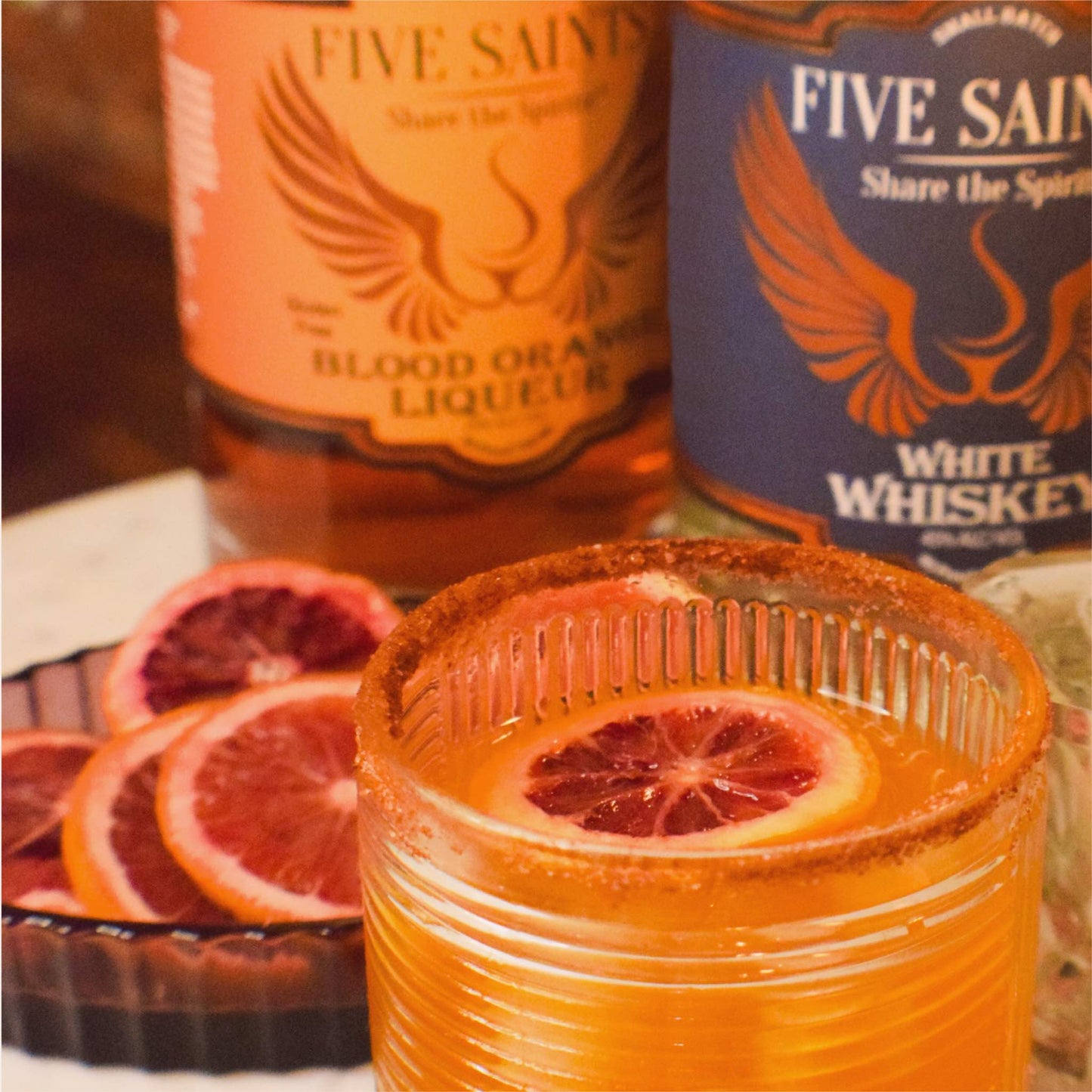 Spicy Confession cocktail in a rocks glass with blood orange garnish, served alongside Five Saints White Whiskey and Blood Orange Liqueur bottles