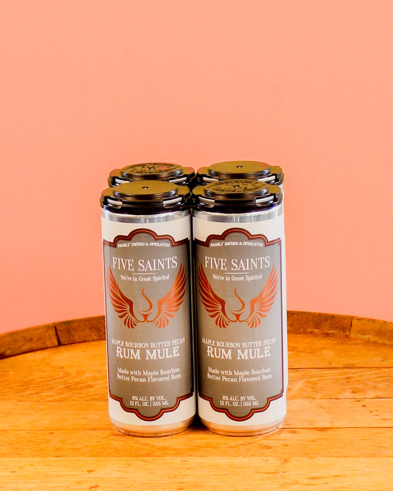 Four bottles of Five Saints Rum Mule canned cocktails on a wooden surface against a peach-colored background.