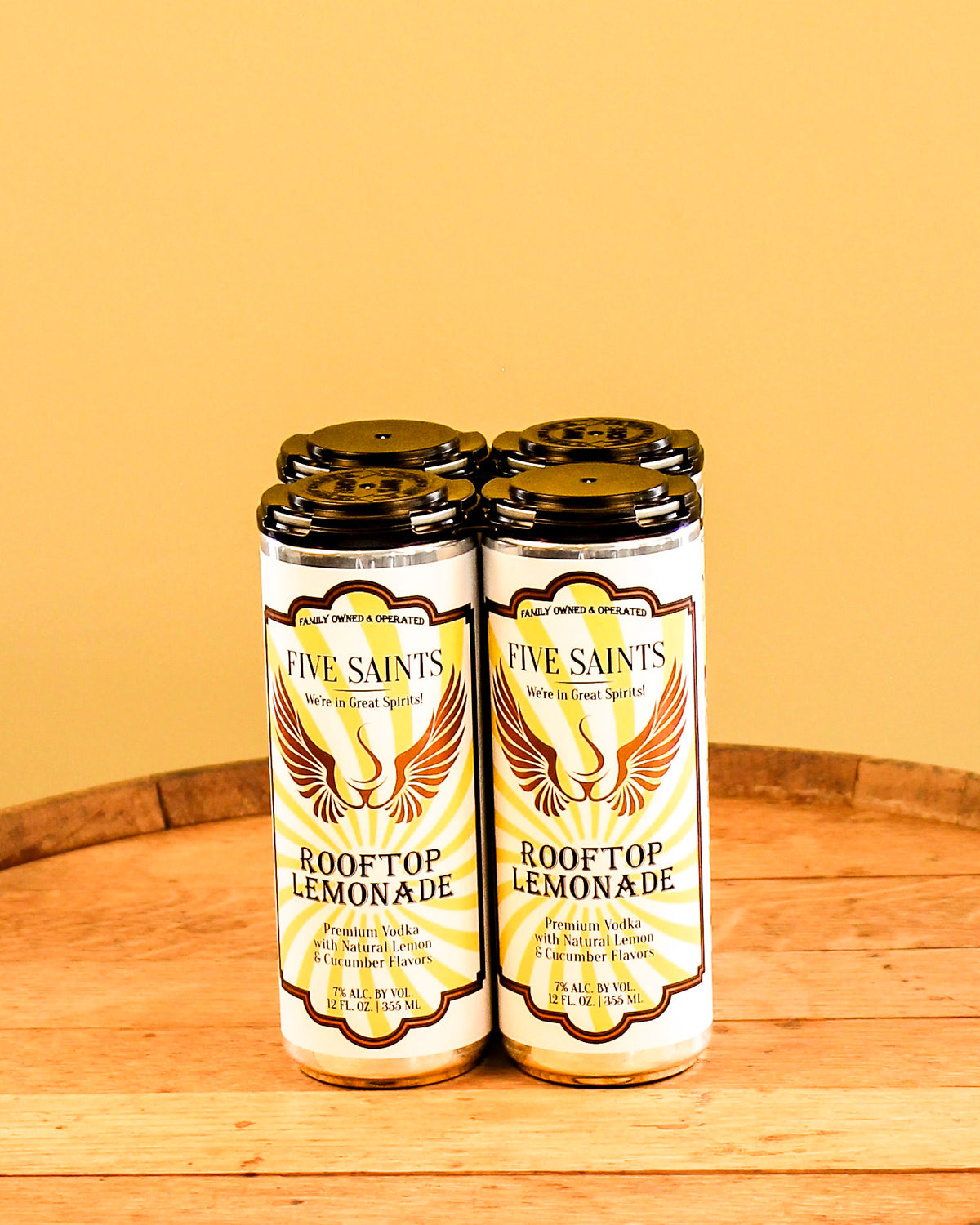 Four cans of Five Saints Rooftop Lemonade arranged side by side on a wooden surface against a warm yellow background.