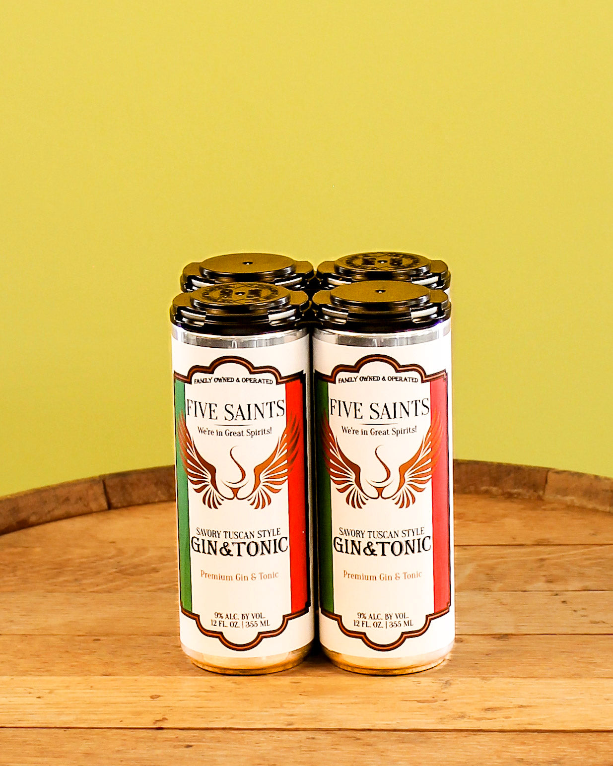 Four bottles of Five Saints Savory Tuscan Style Gin & Tonic canned cocktails on a wooden surface against a yellow-green background.