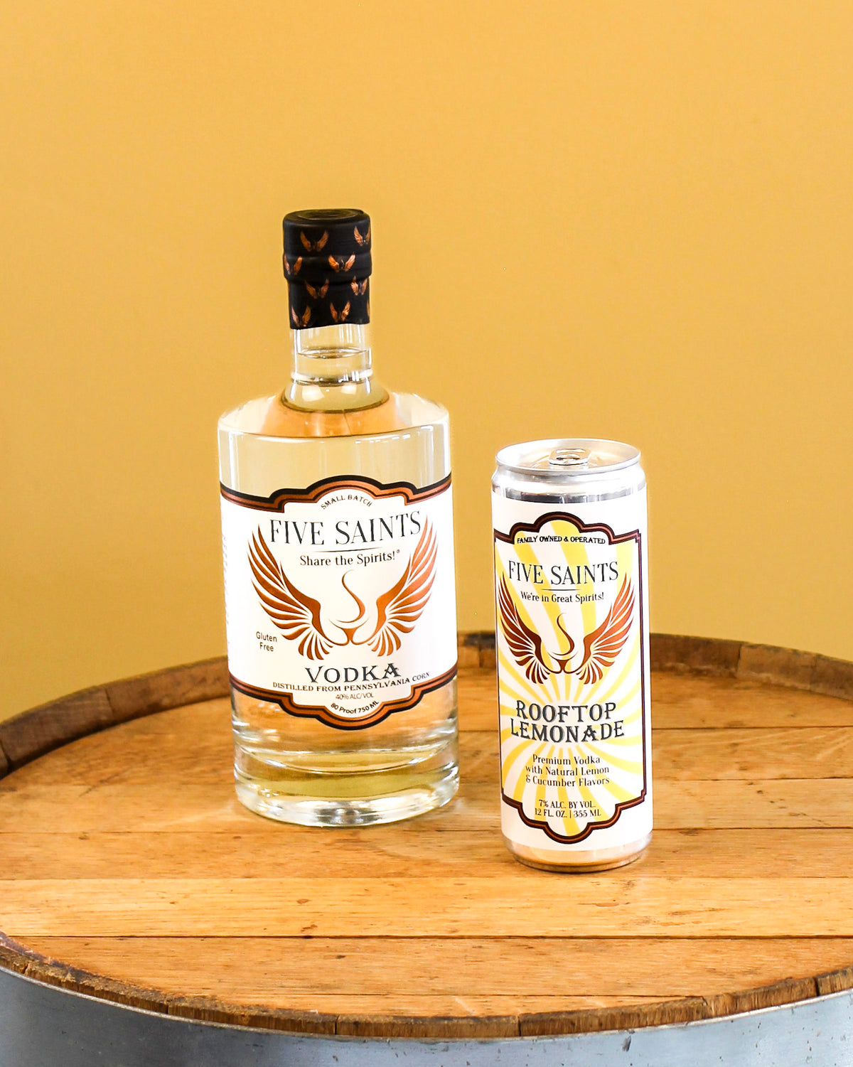 A bottle of Five Saints Vodka and a can of Five Saints Rooftop Lemonade displayed side by side on a wooden barrel top against a solid yellow background.