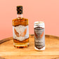 A bottle of Five Saints Humane No.1 Rum and a can of Five Saints Rum Mule side by side on a wooden barrel top against a solid peach-pink background.