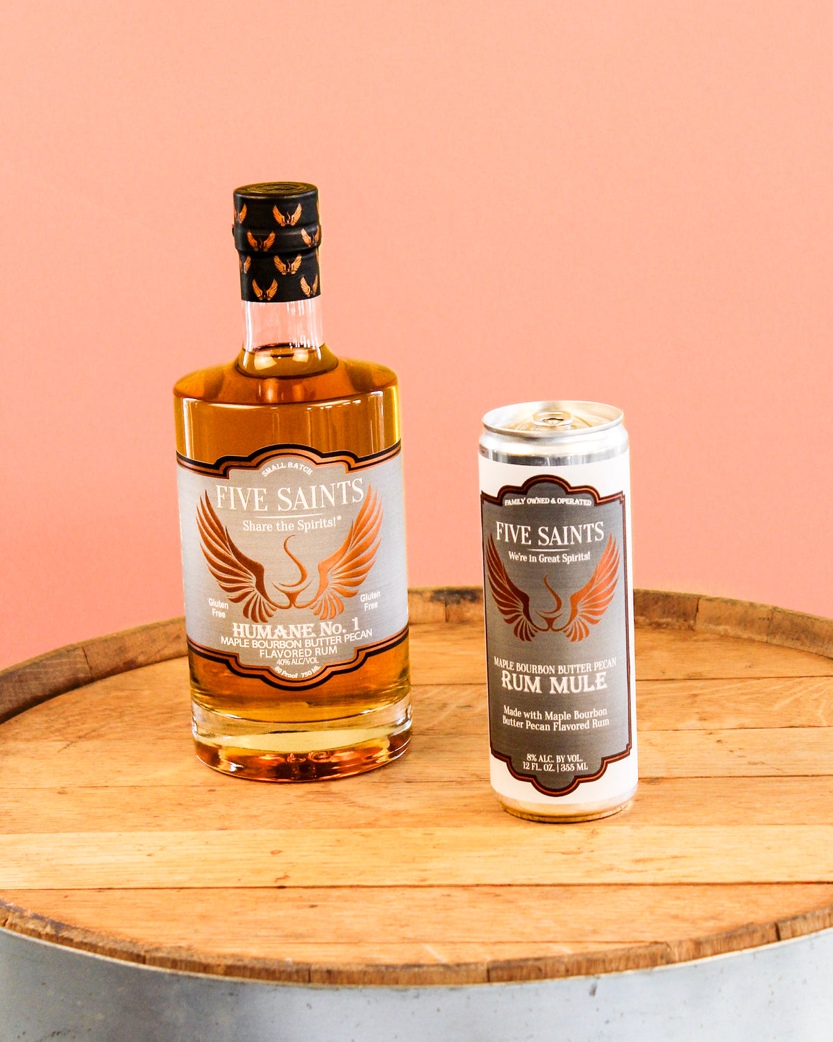 A bottle of Five Saints Humane No.1 Rum and a can of Five Saints Rum Mule side by side on a wooden barrel top against a solid peach-pink background.