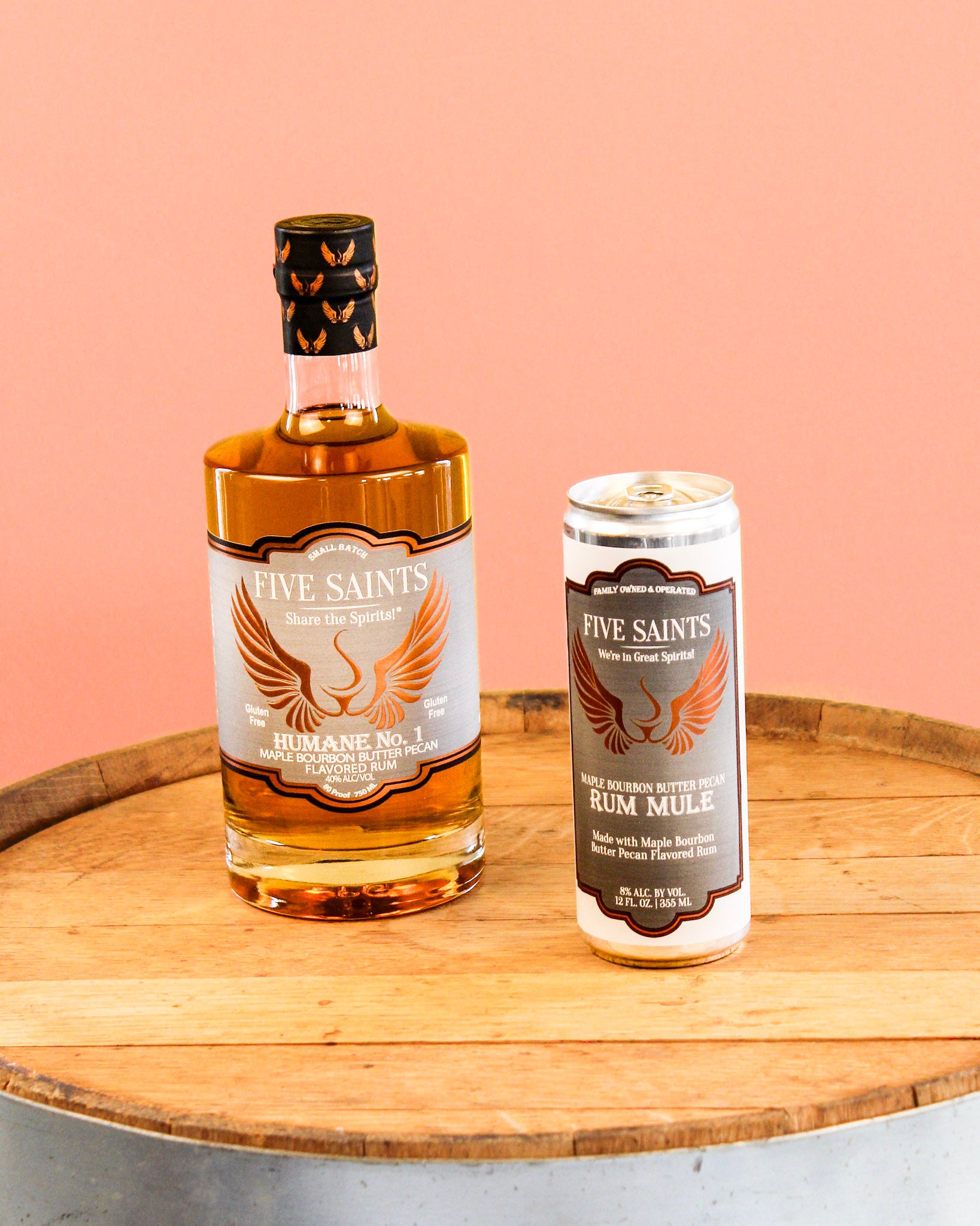 A bottle of Five Saints Humane No.1 Rum and a can of Five Saints Rum Mule side by side on a wooden barrel top against a solid peach-pink background.