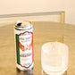 A can of Five Saints Gin & Tonic standing on a reflective metallic table next to a clear ribbed glass filled with ice, against a softly blurred indoor background.