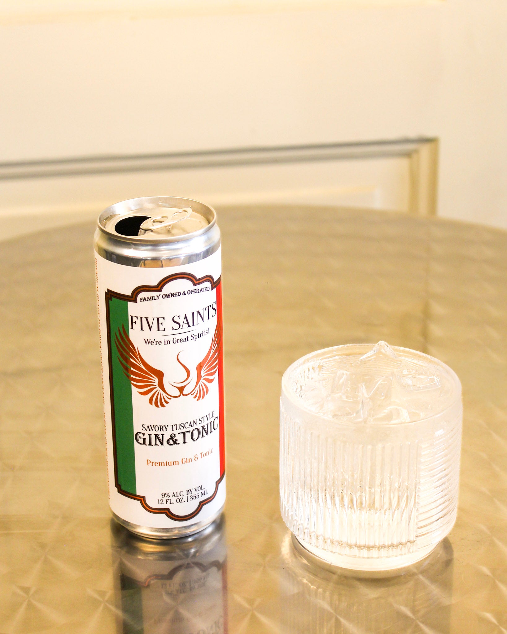 A can of Five Saints Gin & Tonic standing on a reflective metallic table next to a clear ribbed glass filled with ice, against a softly blurred indoor background.