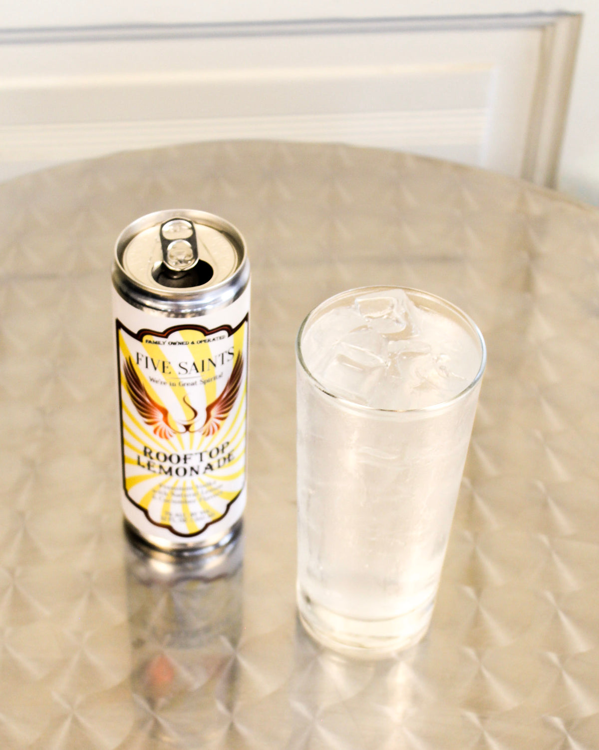 A can of Five Saints Rooftop Lemonade stands next to a clear glass filled with the cocktail stands on a metallic surface