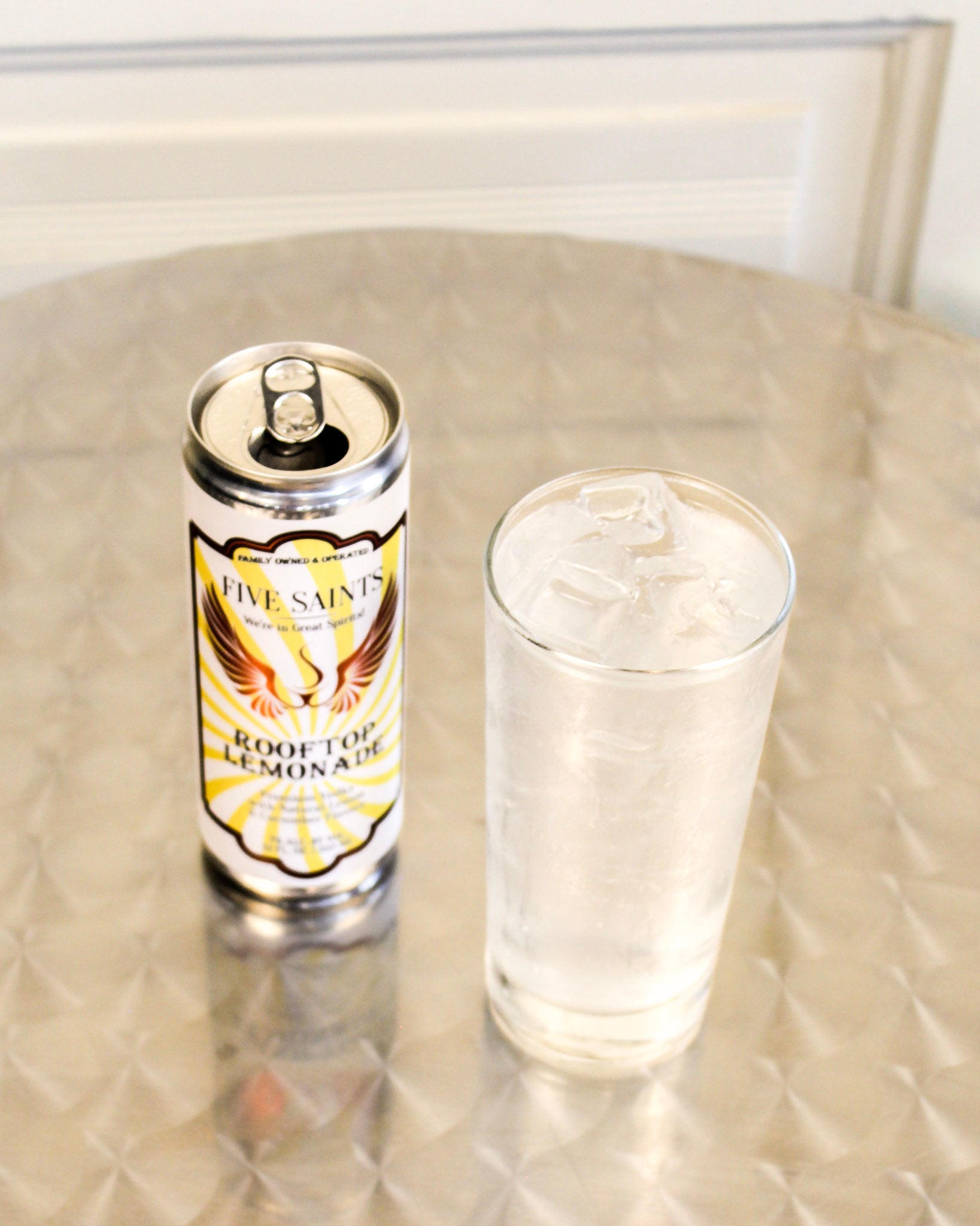 A can of Five Saints Rooftop Lemonade stands next to a clear glass filled with the cocktail stands on a metallic surface