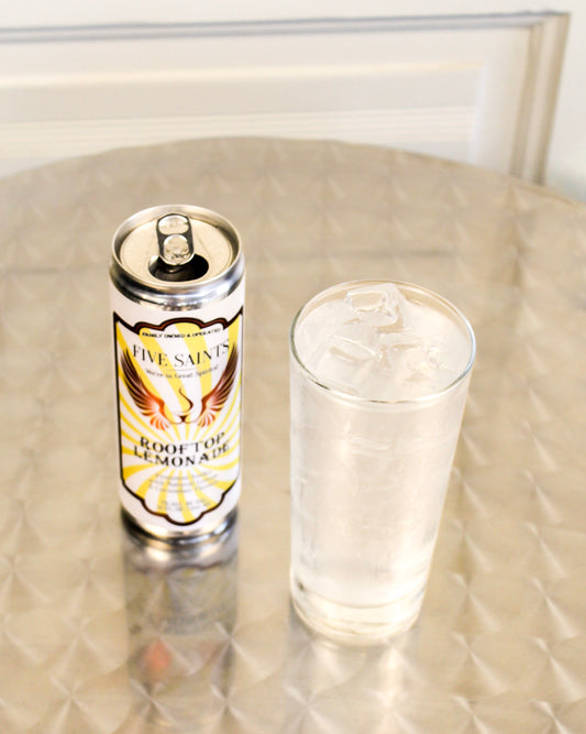 A can of Five Saints Rooftop Lemonade stands next to a clear glass filled with the cocktail stands on a metallic surface