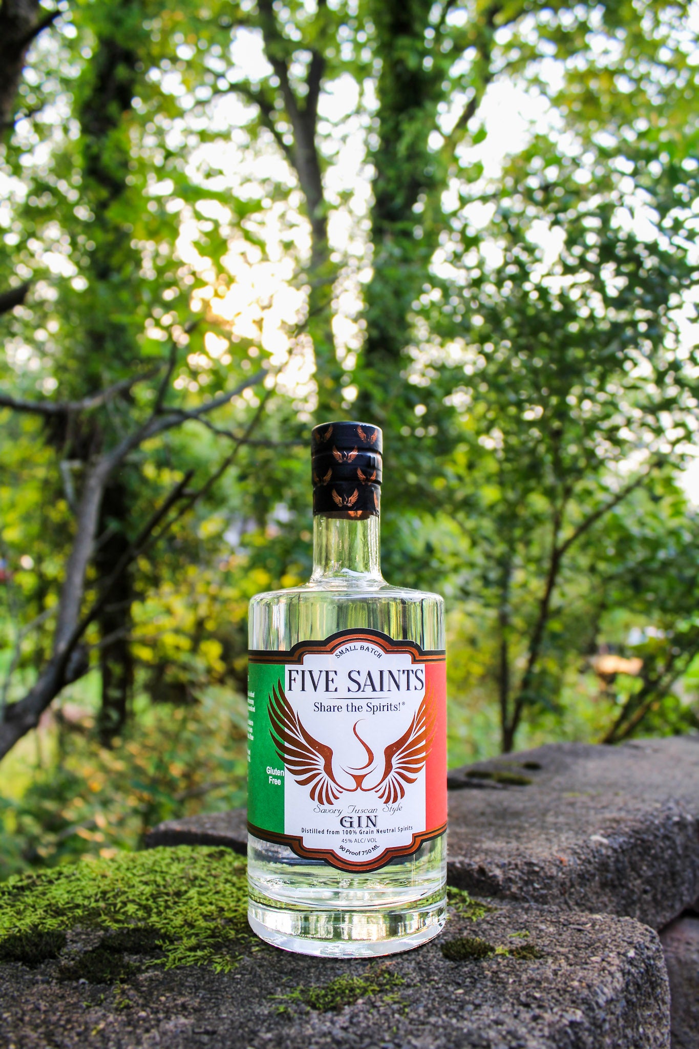 A bottle of Five Saints Gin with a clear glass body and a colorful label, set on a mossy stone with a blurred forest background.