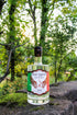 A bottle of Five Saints Gin with a clear glass body and a colorful label, set on a mossy stone with a blurred forest background.
