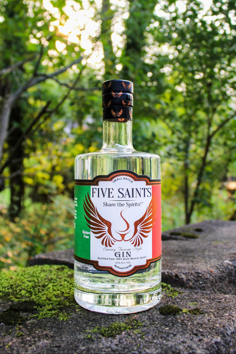 A bottle of Five Saints Gin with a clear glass body and a colorful label, set on a mossy stone with a blurred forest background.